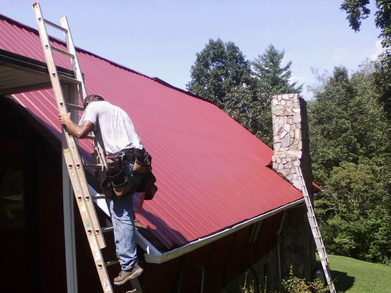 Roofer on a ladder beside a red metal roof and stone chimney — Soffit & Fascia Repair by Roofing Brothers in Rohrersville